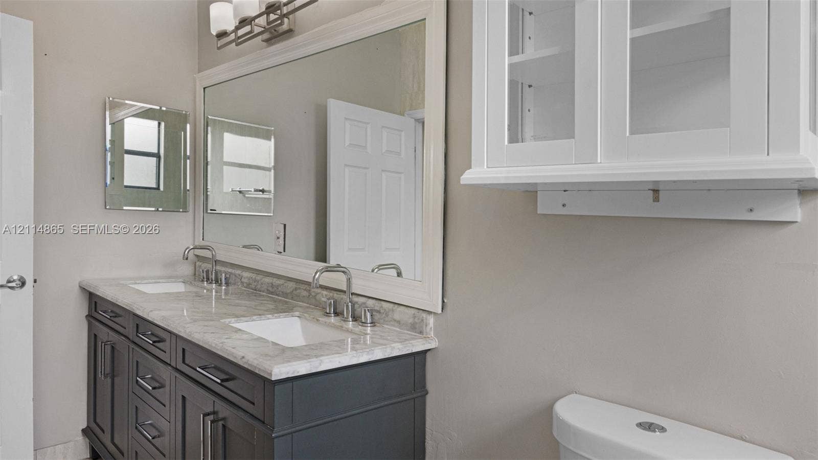 3754 NE 2nd Street, Homestead, FL 33033 Dual-sink vanity with stone-finish countertop, undermount basins, wall-mounted light fixture, large framed mirror, and white built-in upper cabinetry with glass-front doors