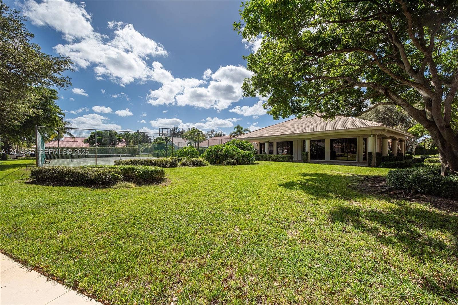 5722 Aspen Ridge Circle, Delray Beach, FL 33484 Expansive rear grounds featuring a fenced tennis court, manicured lawn, mature shade trees, and a covered patio