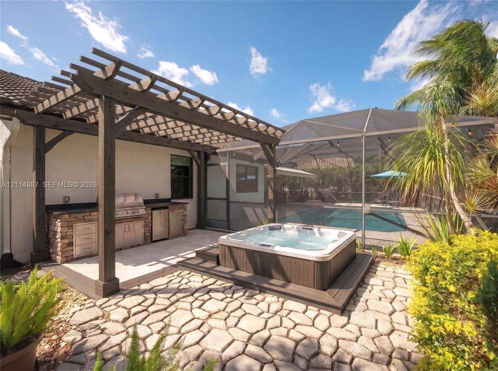 5722 Aspen Ridge Circle, Delray Beach, FL 33484 Outdoor kitchen featuring a stone-clad base, built-in stainless steel grill, and custom cabinetry beneath a dark timber pergola, complemented by an integrated spa and a screened-in swimming pool on a stone paver patio