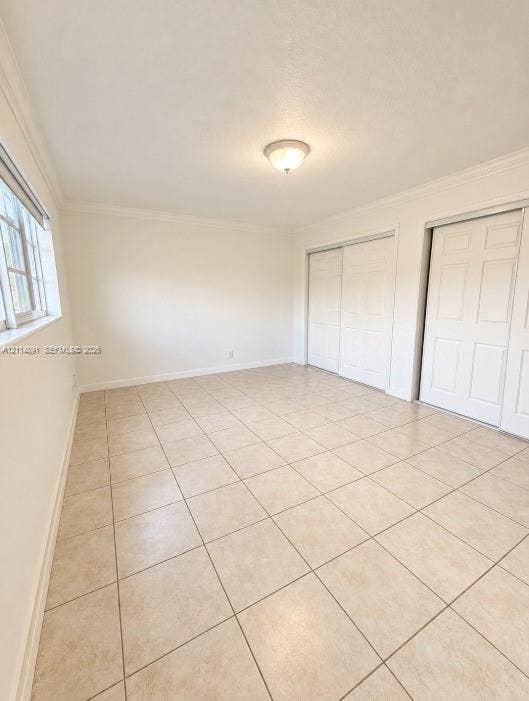 Room featuring neutral tile flooring, crown molding, multi-pane window, and dual sliding-panel closets with six-panel doors