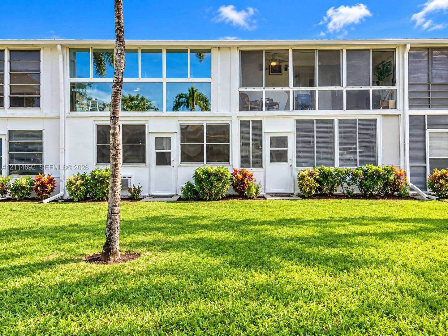 582 Durham U, #582, Deerfield Beach, FL 33442 Multi-level building exterior featuring white stucco facade, expansive floor-to-ceiling windows, screened-in upper-level enclosures, and manicured green space with mature landscaping