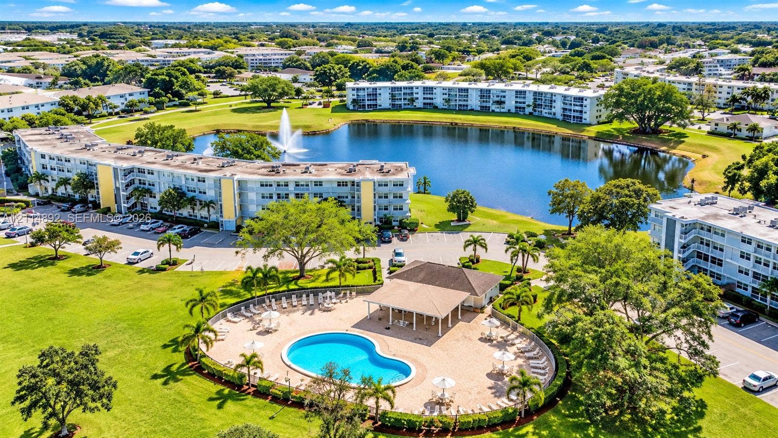 582 Durham U, #582, Deerfield Beach, FL 33442 Aerial perspective of multi-unit residential grounds featuring a swimming pool, covered cabana, expansive pond with a central fountain, and mature landscaping