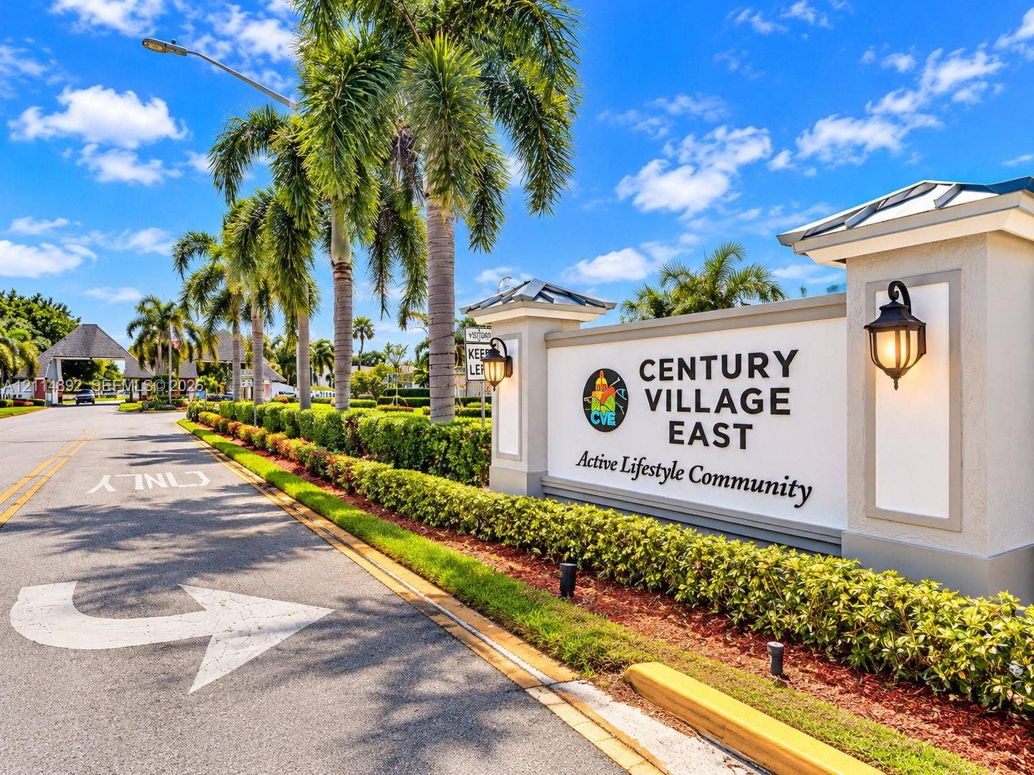 582 Durham U, #582, Deerfield Beach, FL 33442 Community entrance featuring manicured hedges, mature palm trees, decorative wall-mounted lighting, and paved roadway access