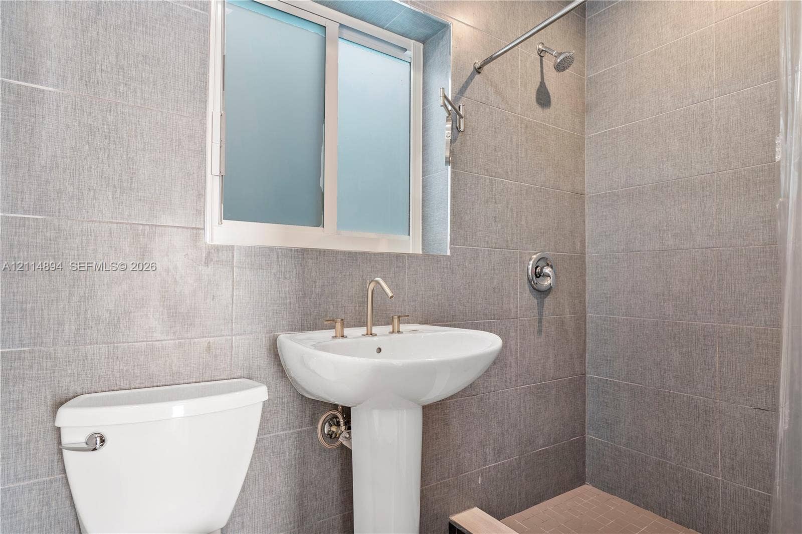Full-height gray textured wall tiling, white pedestal sink with metal widespread faucet, and shower head with integrated control handle