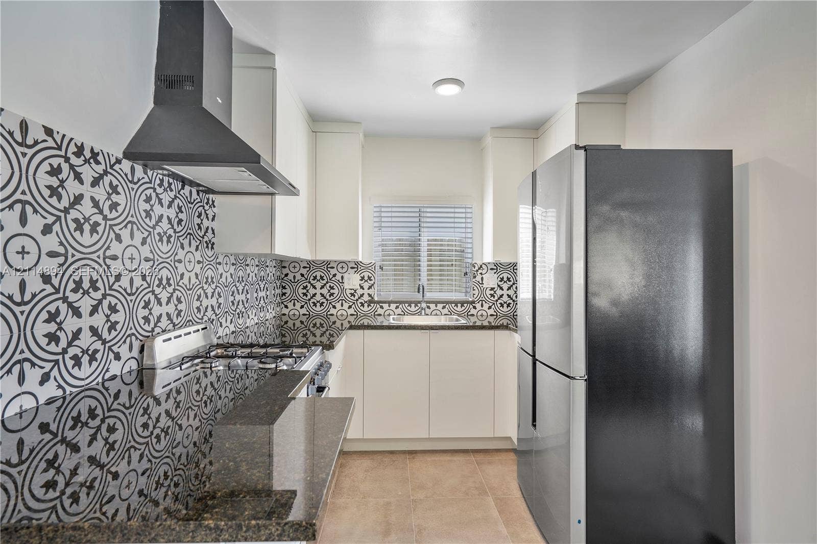 Kitchen featuring patterned tile backsplash, dark stone-finish countertops, matte black range hood, sleek white cabinetry, and stainless steel refrigerator