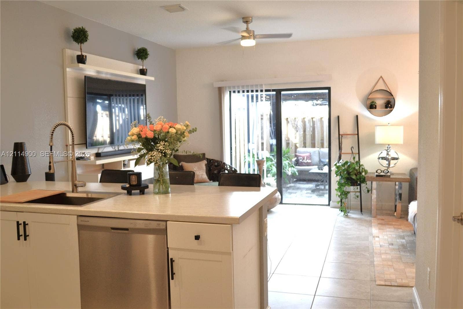 25048 SW 113th Place, Homestead, FL 33032 Kitchen island featuring a stainless steel dishwasher, undermount sink, and high-arc faucet, accompanied by a built-in entertainment center with integrated shelving, tile flooring, and sliding glass patio doors