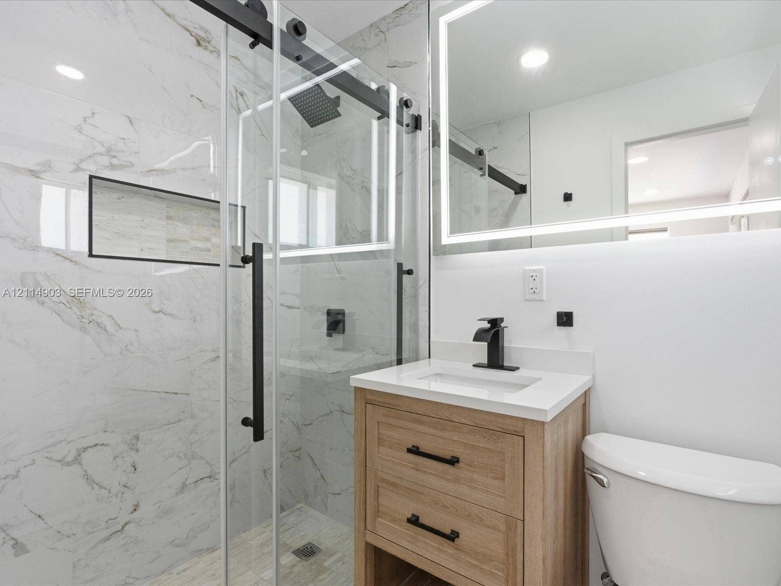 1129 NE 16th Avenue, #1, Fort Lauderdale, Fort Lauderdale, FL 33304 Bathroom featuring a sliding glass shower door with matte black hardware, stone-finish wall tile with integrated recessed shelving, a light wood-finish vanity, a quartz countertop, and an LED-backlit mirror