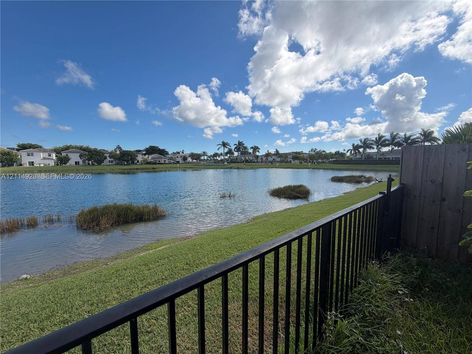 1411 SE 26th Road, Homestead, FL 33035 Waterfront lot featuring a manicured lawn, perimeter metal fencing, and wood-paneled privacy screening