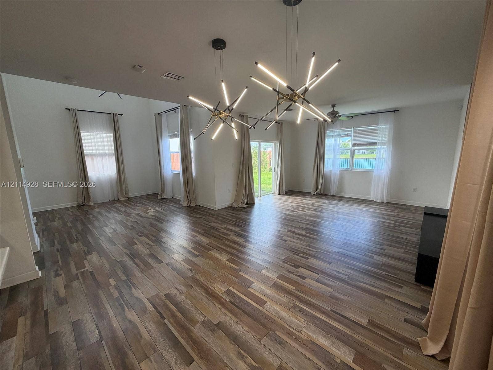 1411 SE 26th Road, Homestead, FL 33035 Open-concept interior featuring wood-finish flooring, high ceilings, dual geometric multi-arm pendant light fixtures, and large sliding glass doors providing access to the outdoors