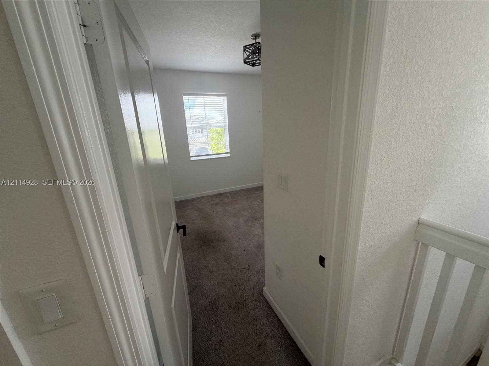 1411 SE 26th Road, Homestead, FL 33035 Interior space featuring neutral-toned carpeting, a geometric metal chandelier, a singular window with horizontal blinds, and a solid-panel door