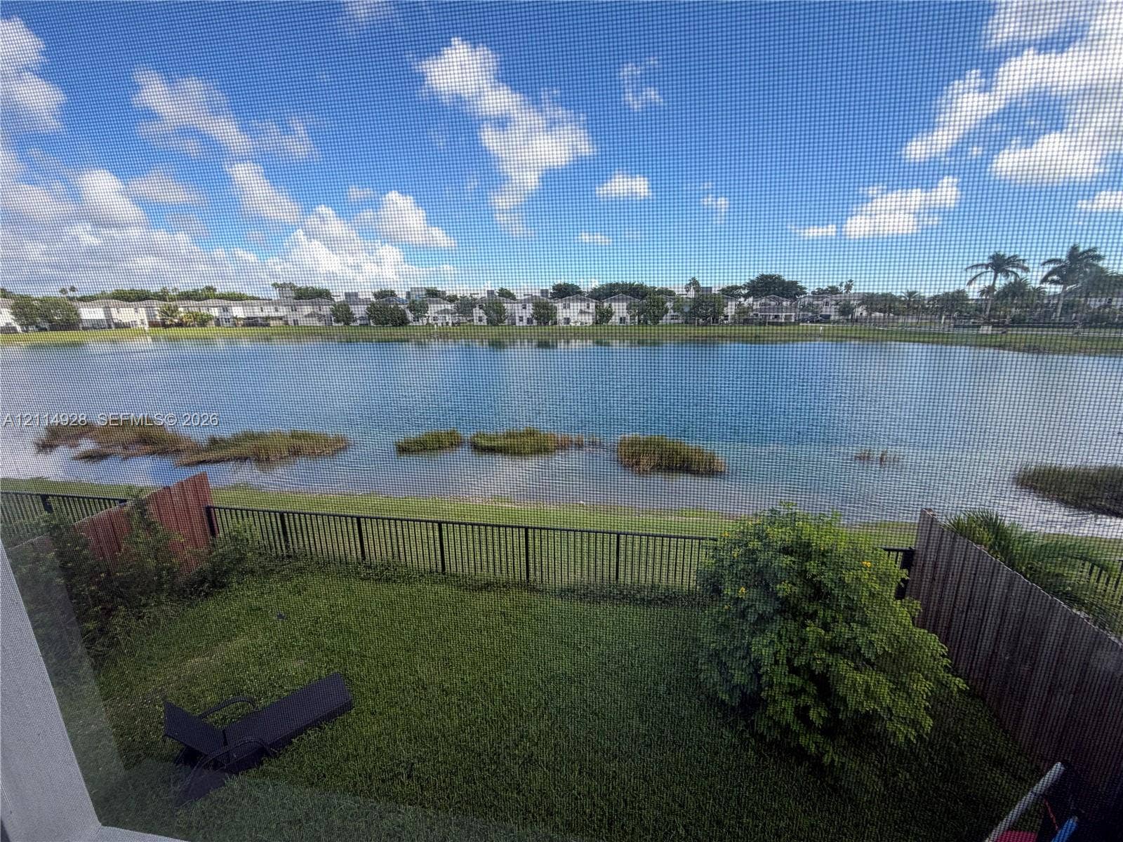 1411 SE 26th Road, Homestead, FL 33035 Waterfront property featuring a grass lawn, perimeter metal fencing, and wood-paneled privacy partitions
