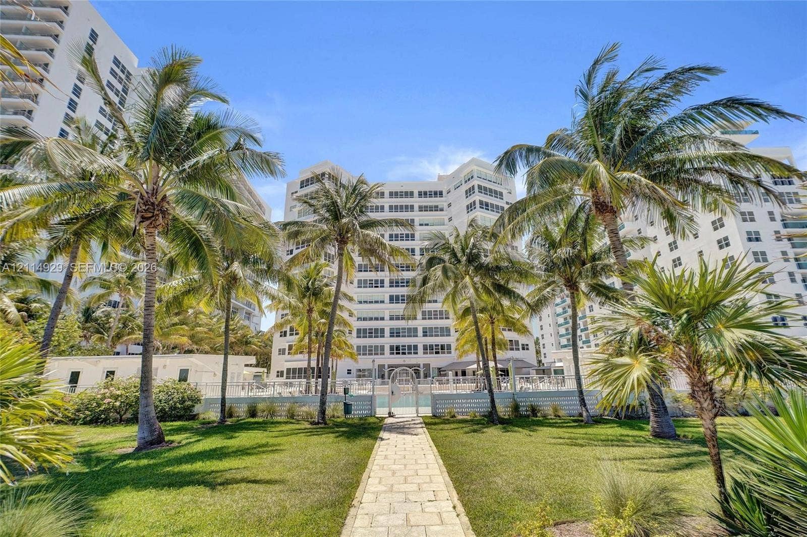 High-rise exterior with white stucco facade, stone-paver walkway, manicured lawn, and mature palm trees