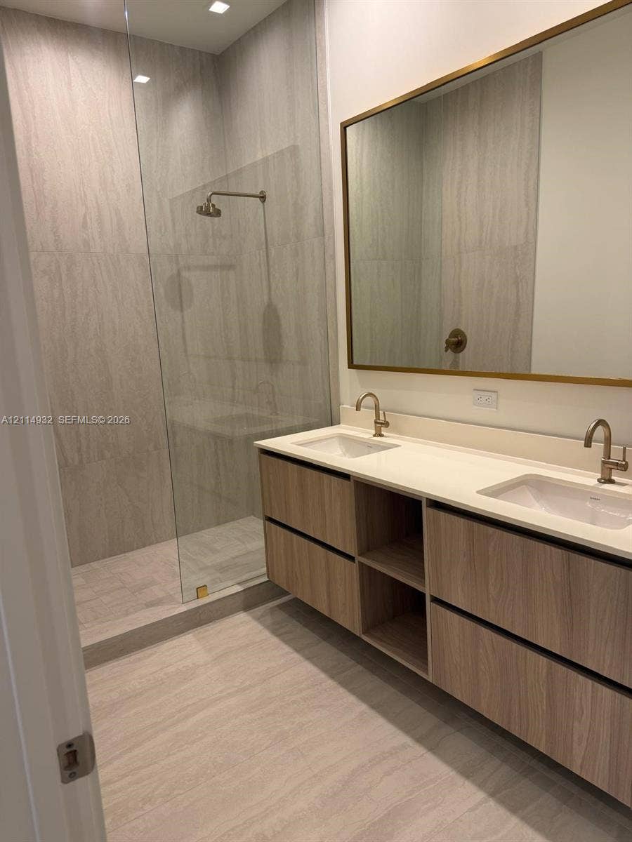 77 SE 5th Street, #607, Miami, FL 33141 Floating dual-vanity with wood-finish cabinetry, white countertop, integrated rectangular sinks, brass-toned fixtures, and a glass-enclosed shower with floor-to-ceiling tile