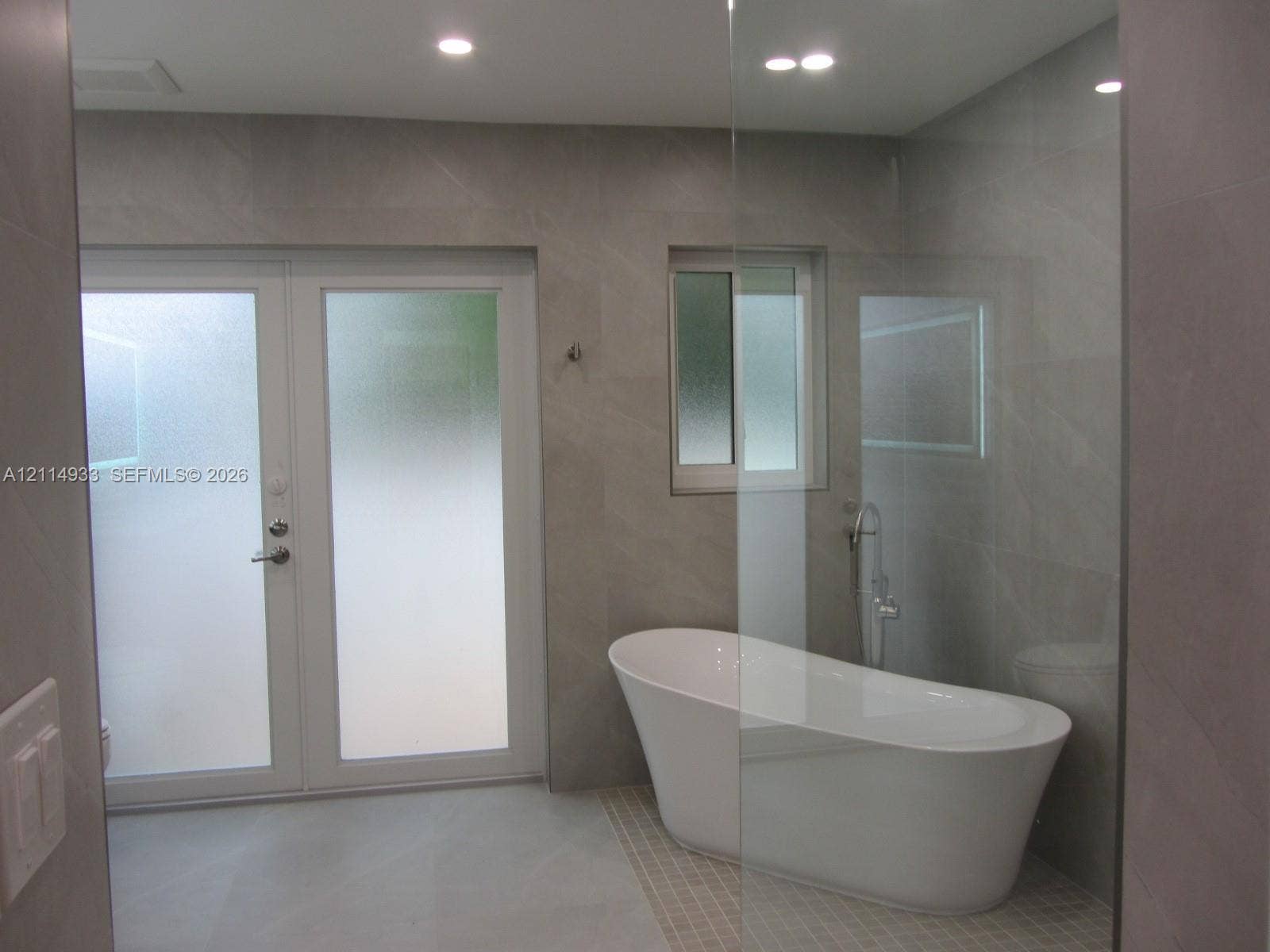 1001 Bella Vista Avenue, Coral Gables, Coral Gables, FL 33156 Freestanding soaking tub with a floor-mounted faucet, large-format grey tile wall cladding, glass shower enclosure, and white frosted glass double doors