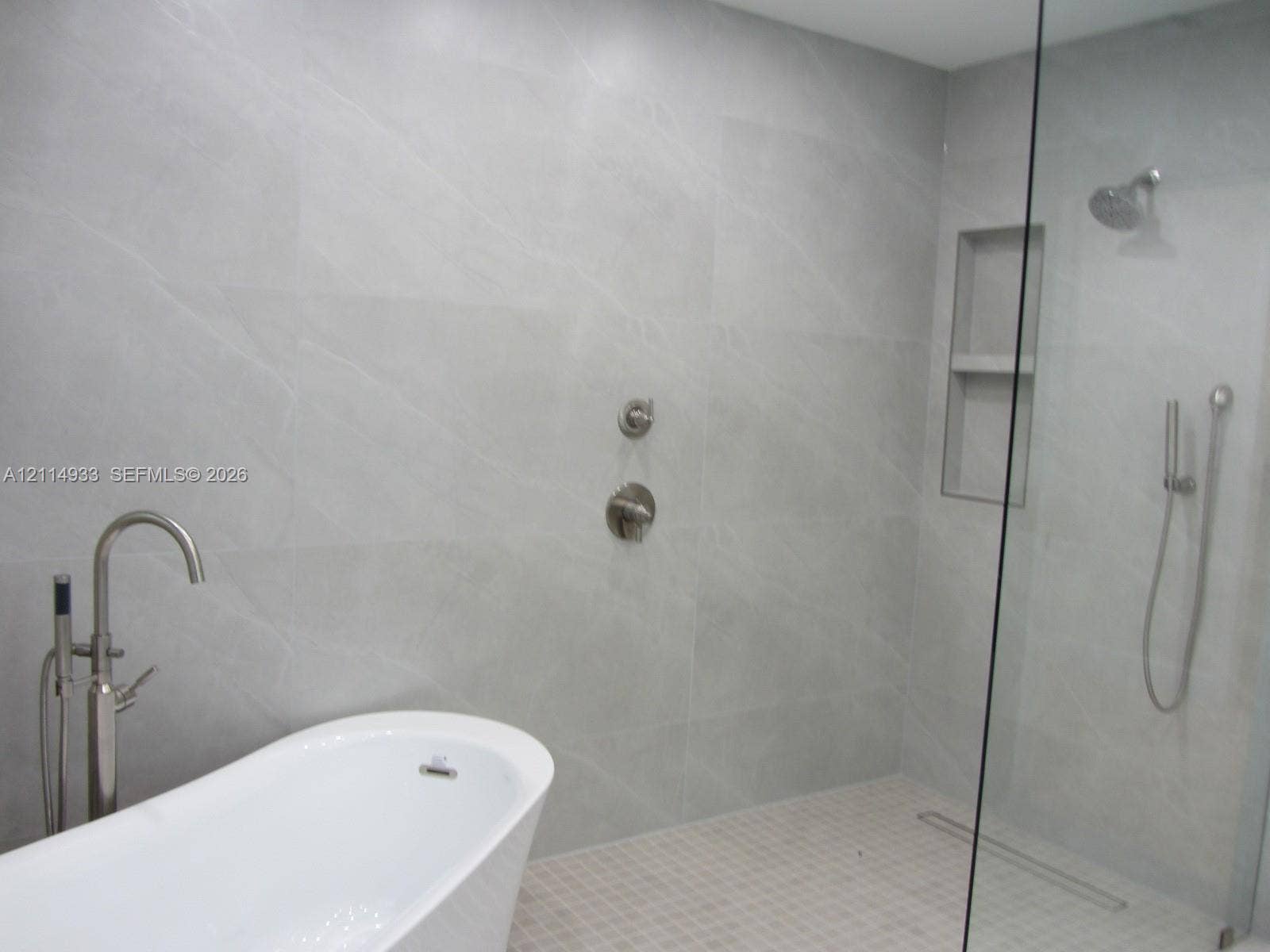 1001 Bella Vista Avenue, Coral Gables, Coral Gables, FL 33156 Freestanding soaking tub with floor-mounted faucet, walk-in shower featuring large-format tile surround, integrated tiled wall niche, rain shower head with handheld attachment, and seamless glass partition
