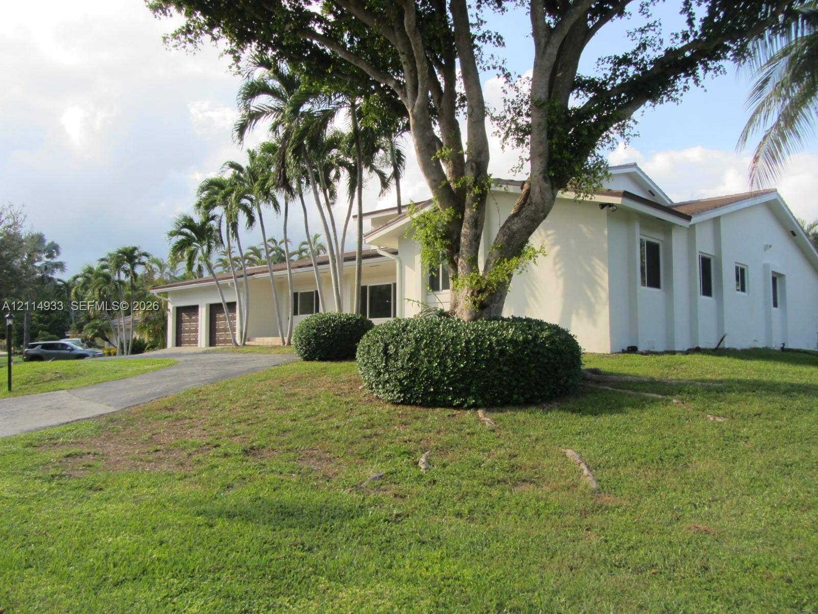 1001 Bella Vista Avenue, Coral Gables, Coral Gables, FL 33156 Stucco exterior featuring a two-car garage, expansive lawn, mature shade trees, and ornamental palms