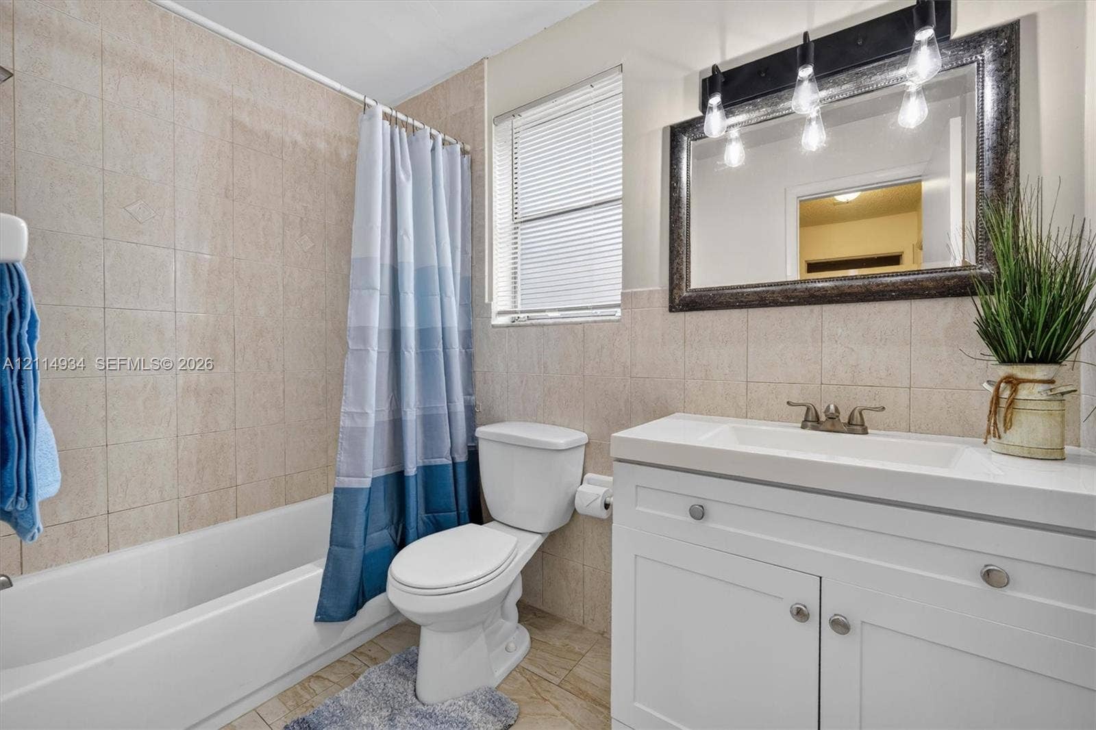 Bathroom featuring floor-to-ceiling neutral wall tile, white shaker-style vanity with an integrated sink, three-light wall-mounted fixture, and a framed mirror