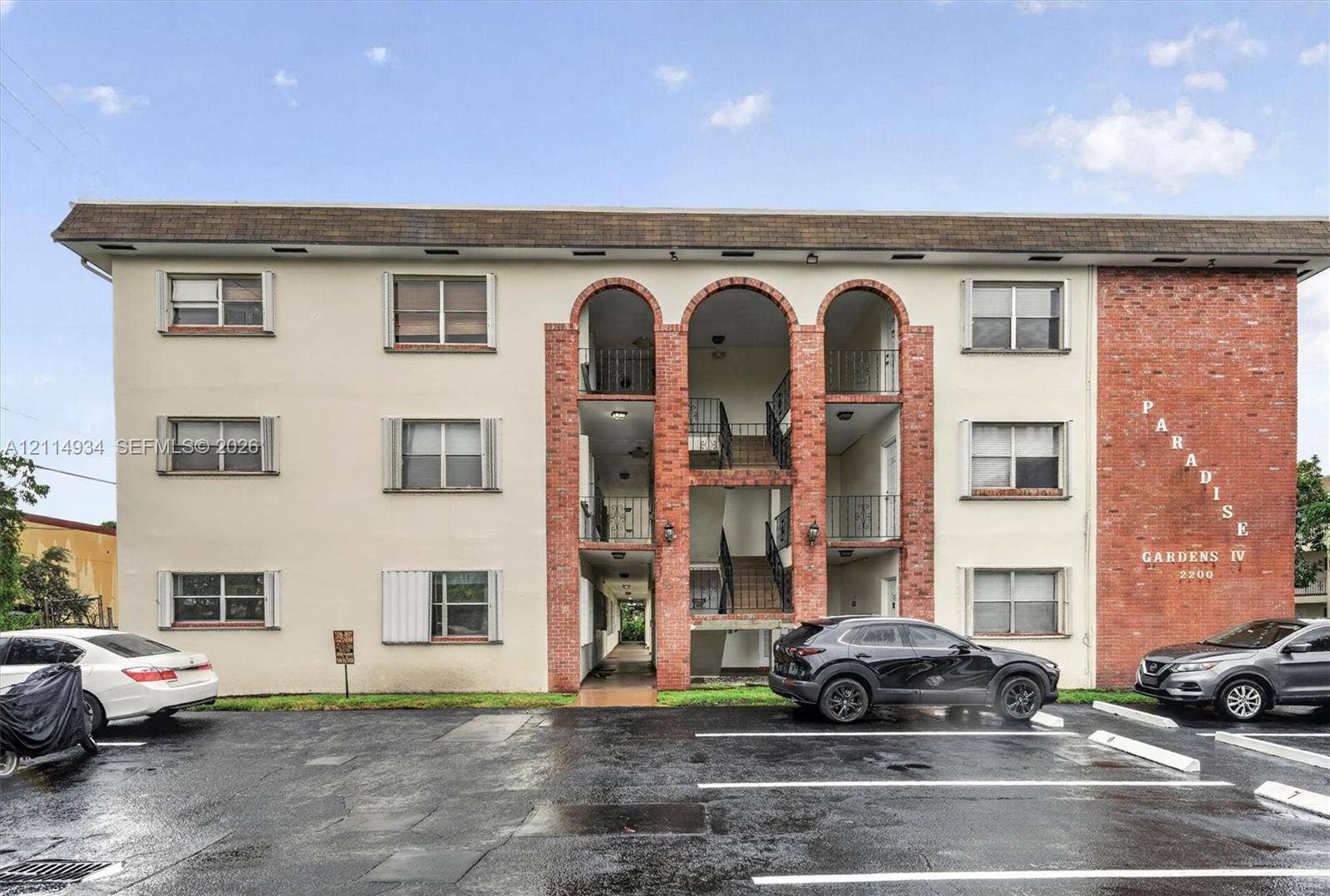 Exterior facade featuring a combination of stucco and red brick masonry, arched entryway detailing, metal balcony railings, and surface parking