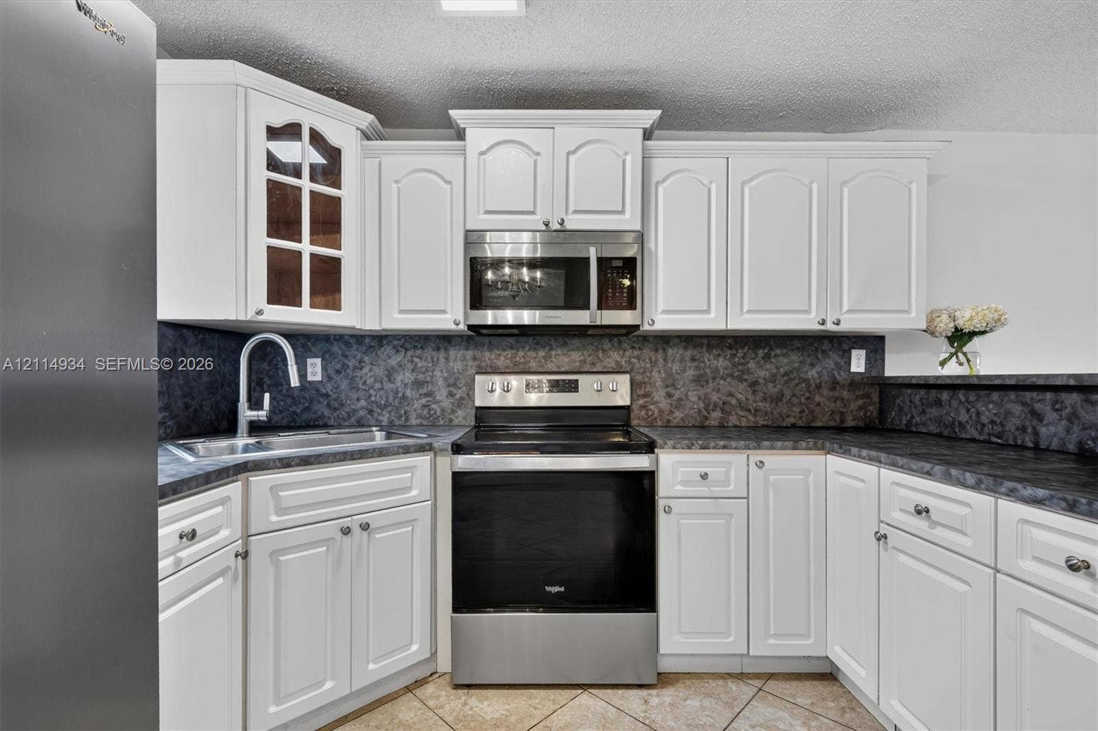 Kitchen featuring white cabinetry with glass-panel accents, dark stone-patterned countertops, stainless steel appliances, a high-arc faucet, and tiled flooring