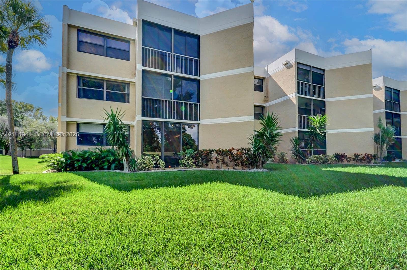 16175 Golf Club Road, #113, Weston, FL 33326 Multi-story exterior featuring a neutral stucco facade, stacked balconies with metal railings, expansive glass windows, and mature perimeter landscaping