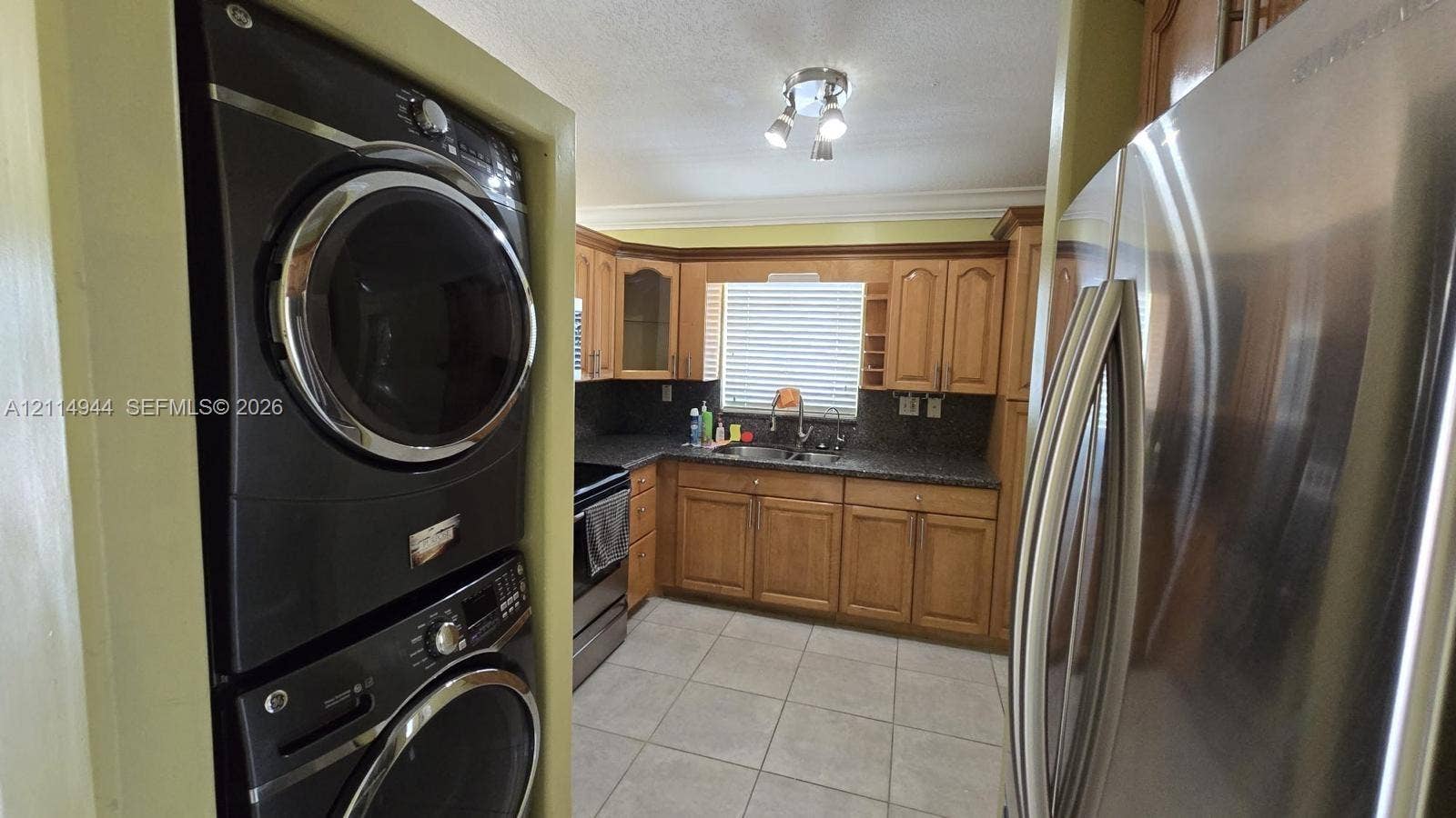 Kitchen featuring medium-tone wood cabinetry, dark stone-finish countertops, stainless steel side-by-side refrigerator, stackable laundry unit, and ceramic tile flooring