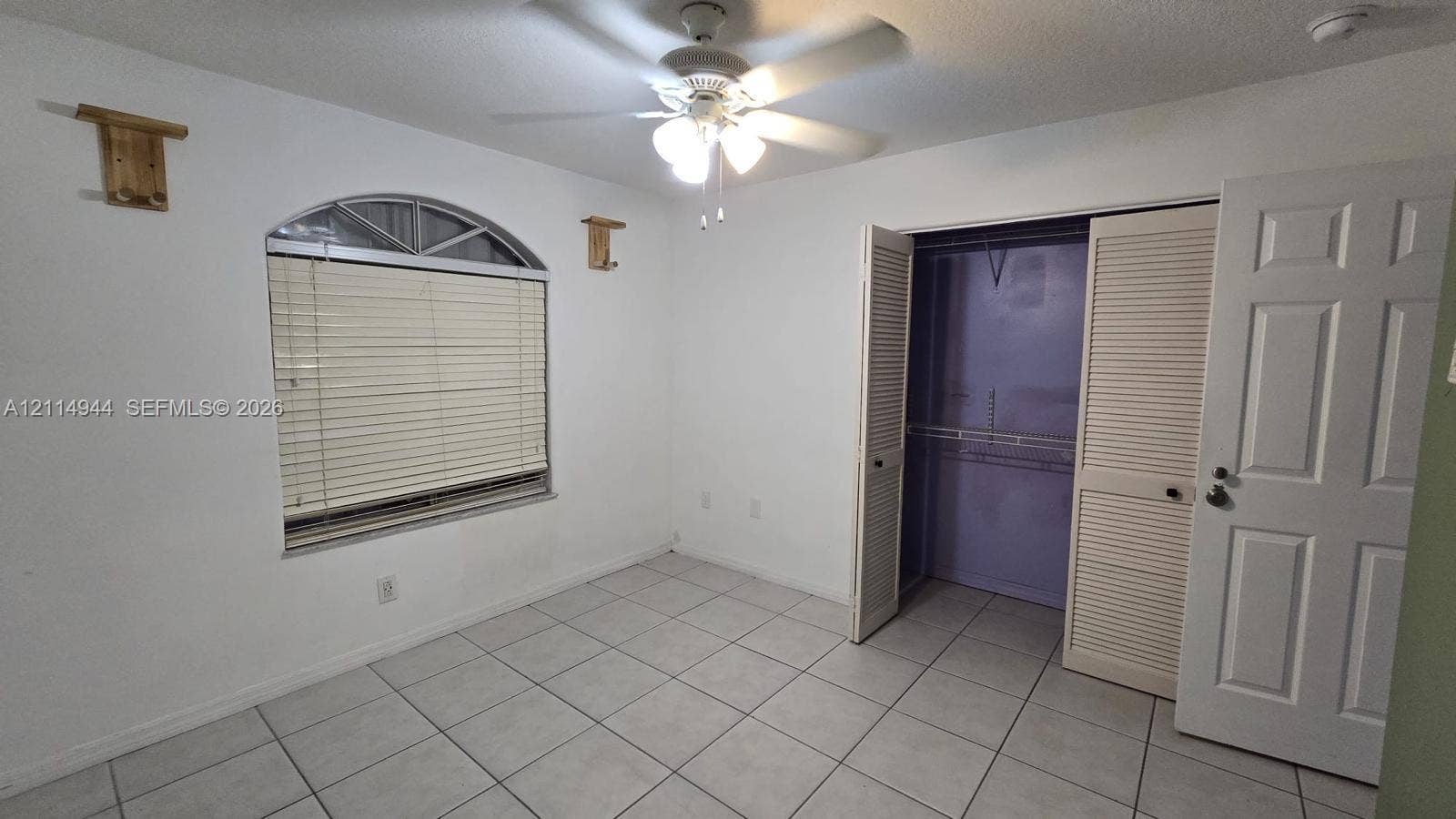 Room featuring large-format tile flooring, arched window with a fanlight, ceiling fan with light fixture, and a closet with louvered bifold doors