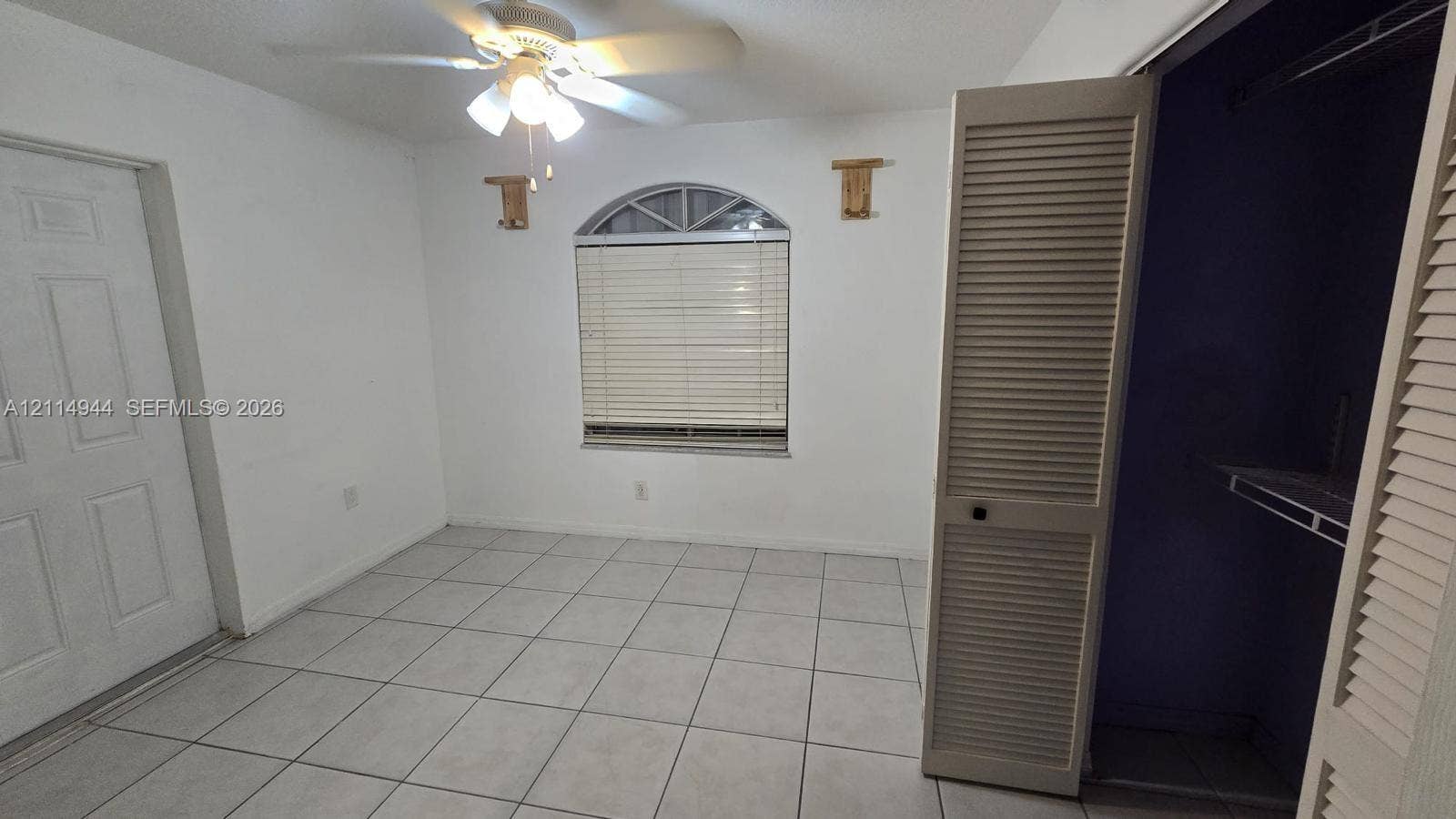 Interior space featuring ceramic tile flooring, arched window with horizontal blinds, ceiling fan with integrated lighting, and louvered closet doors