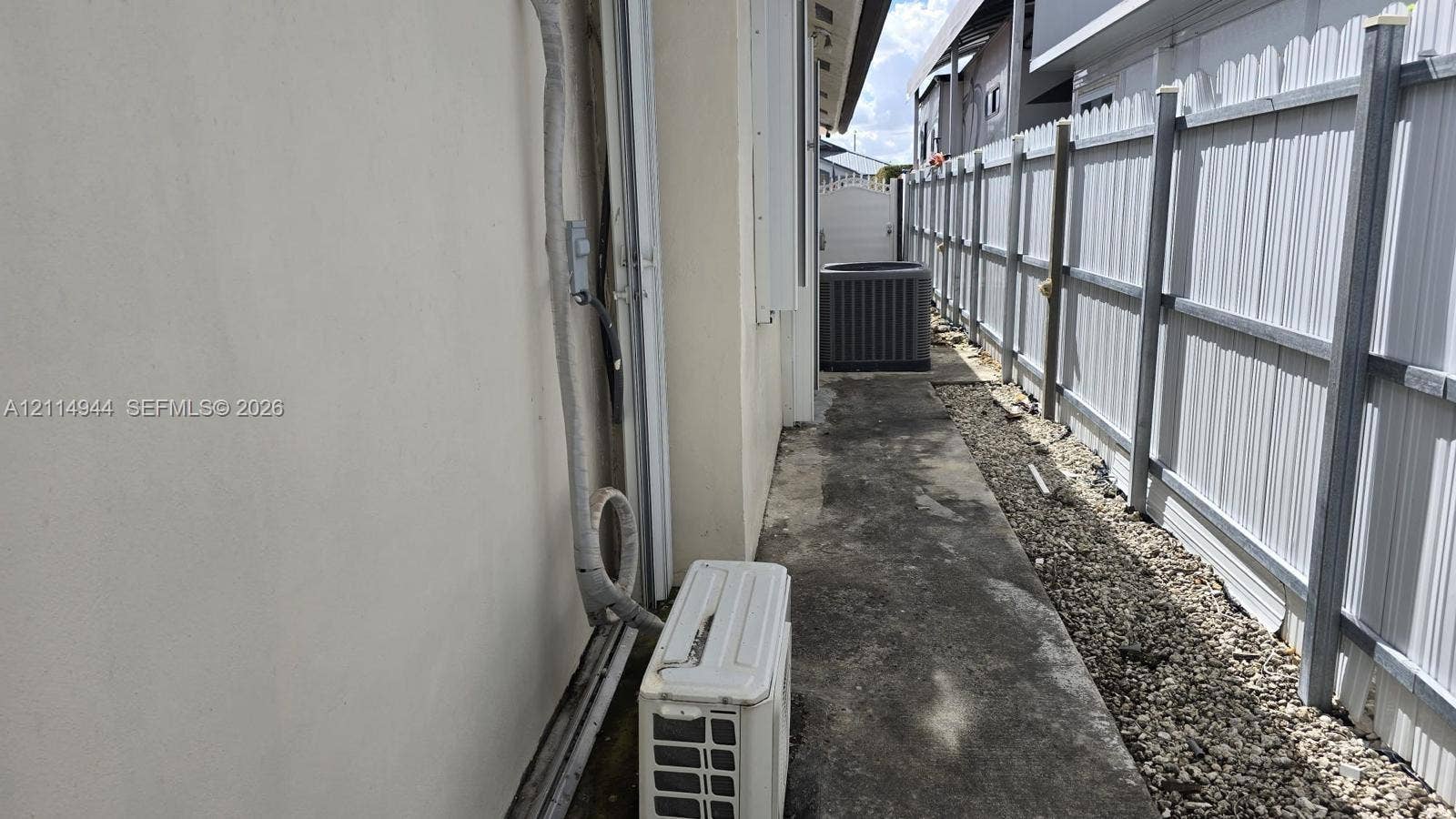 Side exterior featuring a concrete walkway, light-colored stucco siding, a metal privacy fence, and an exterior air conditioning unit
