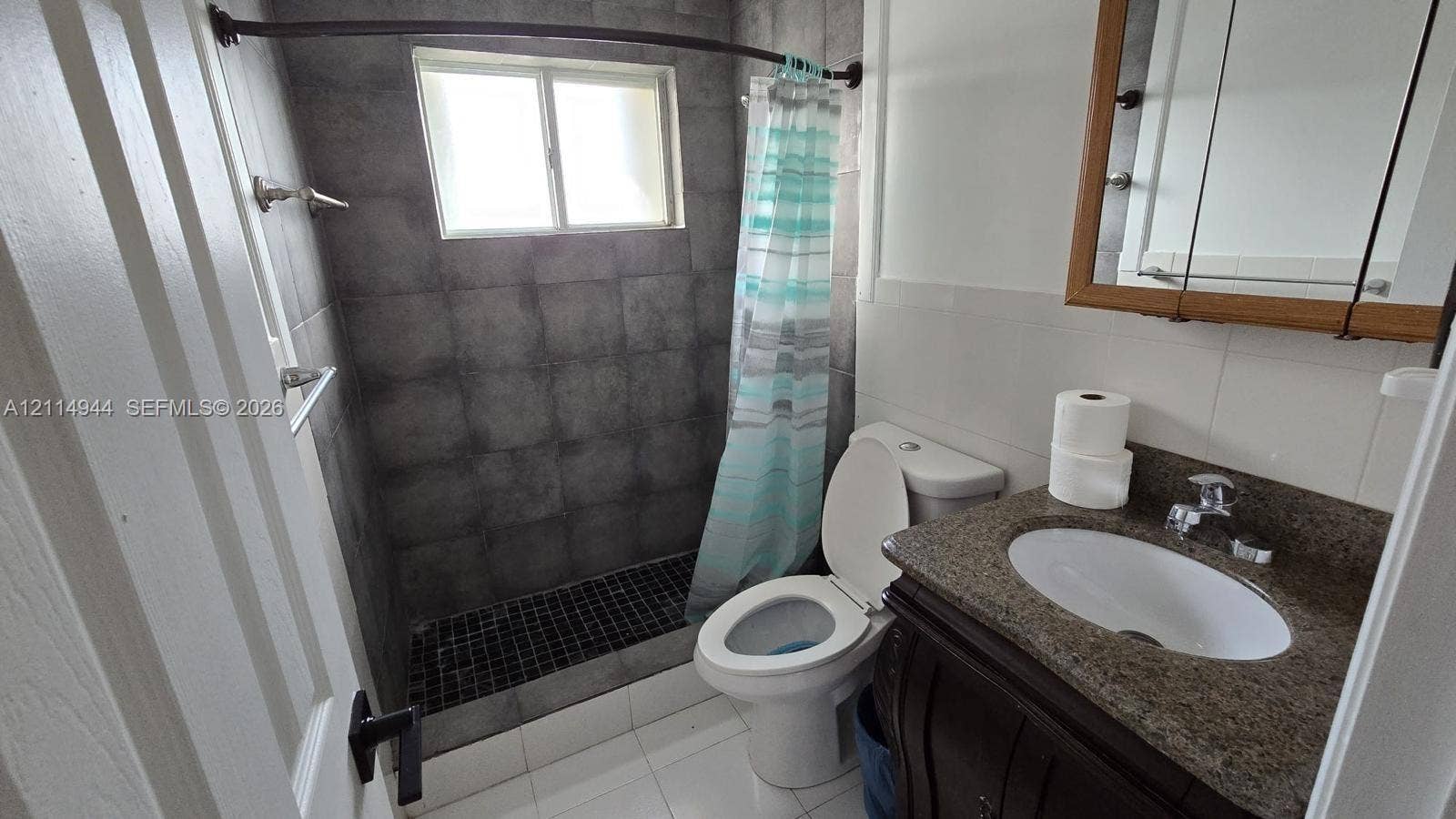 Bathroom featuring a walk-in shower with charcoal-toned wall tiles, dark mosaic floor tiling, stone-surfaced vanity with an integrated basin, and a wood-finish medicine cabinet