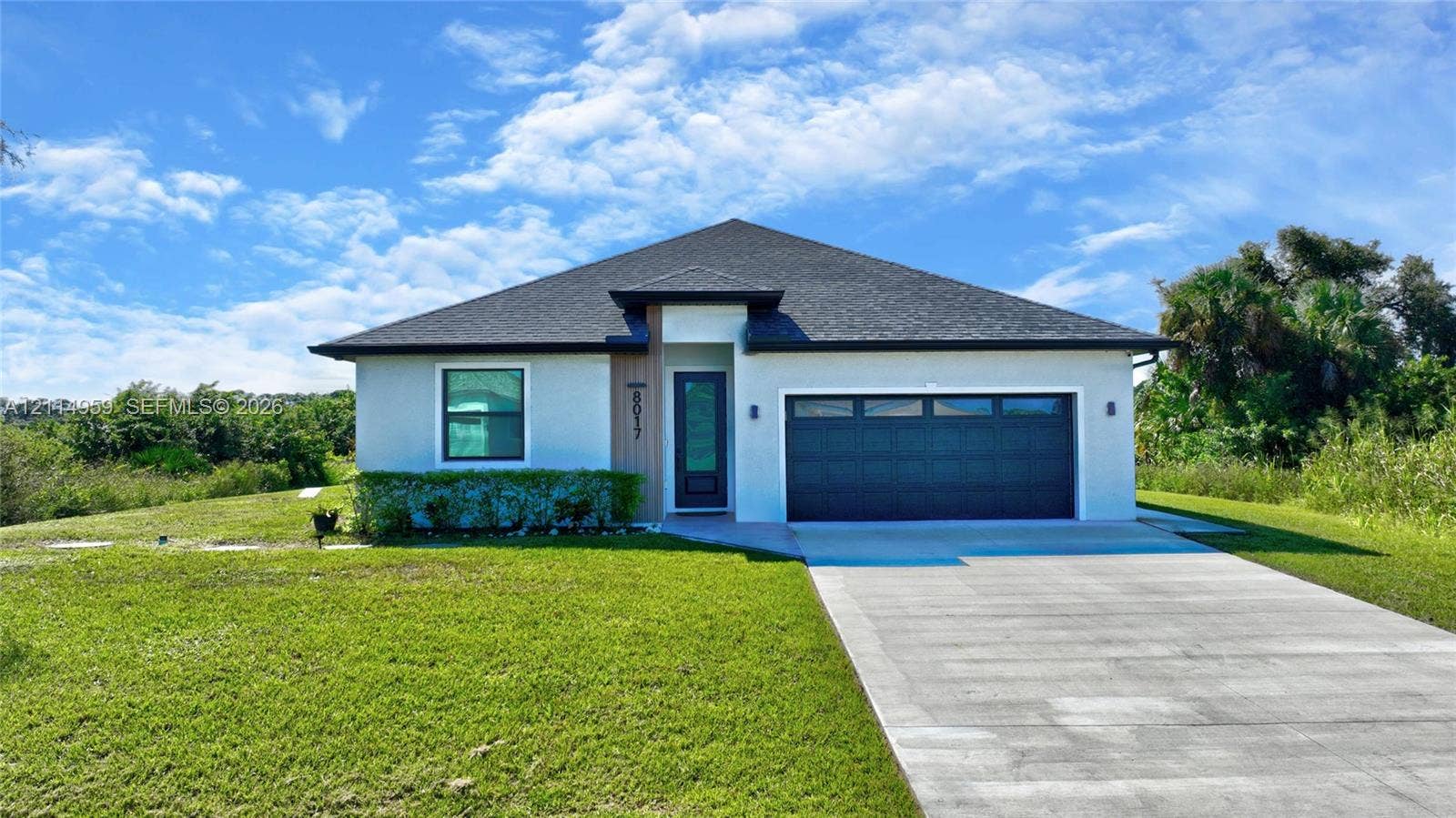 8017 Sunrise Circle, Labelle, FL 33935 Single-story exterior featuring a light-toned stucco faade, wood-finish entryway accent, attached two-car garage, and a hip roofline