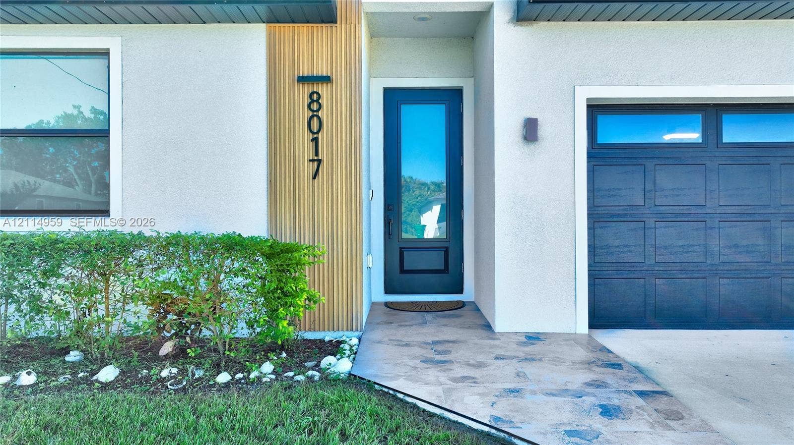 8017 Sunrise Circle, Labelle, FL 33935 Modern entryway featuring a vertical wood-slat accent, black-framed glass panel door, stone-finish walkway, and multi-pane garage door