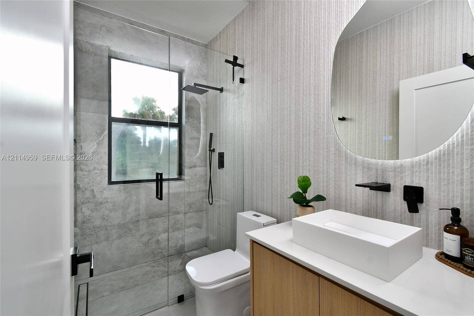 8017 Sunrise Circle, Labelle, FL 33935 Bathroom featuring a floating wood-finish vanity with a rectangular vessel sink, wall-mounted matte black faucet fixtures, a frameless glass shower enclosure with large-format tile, and an illuminated organic-shaped mirror