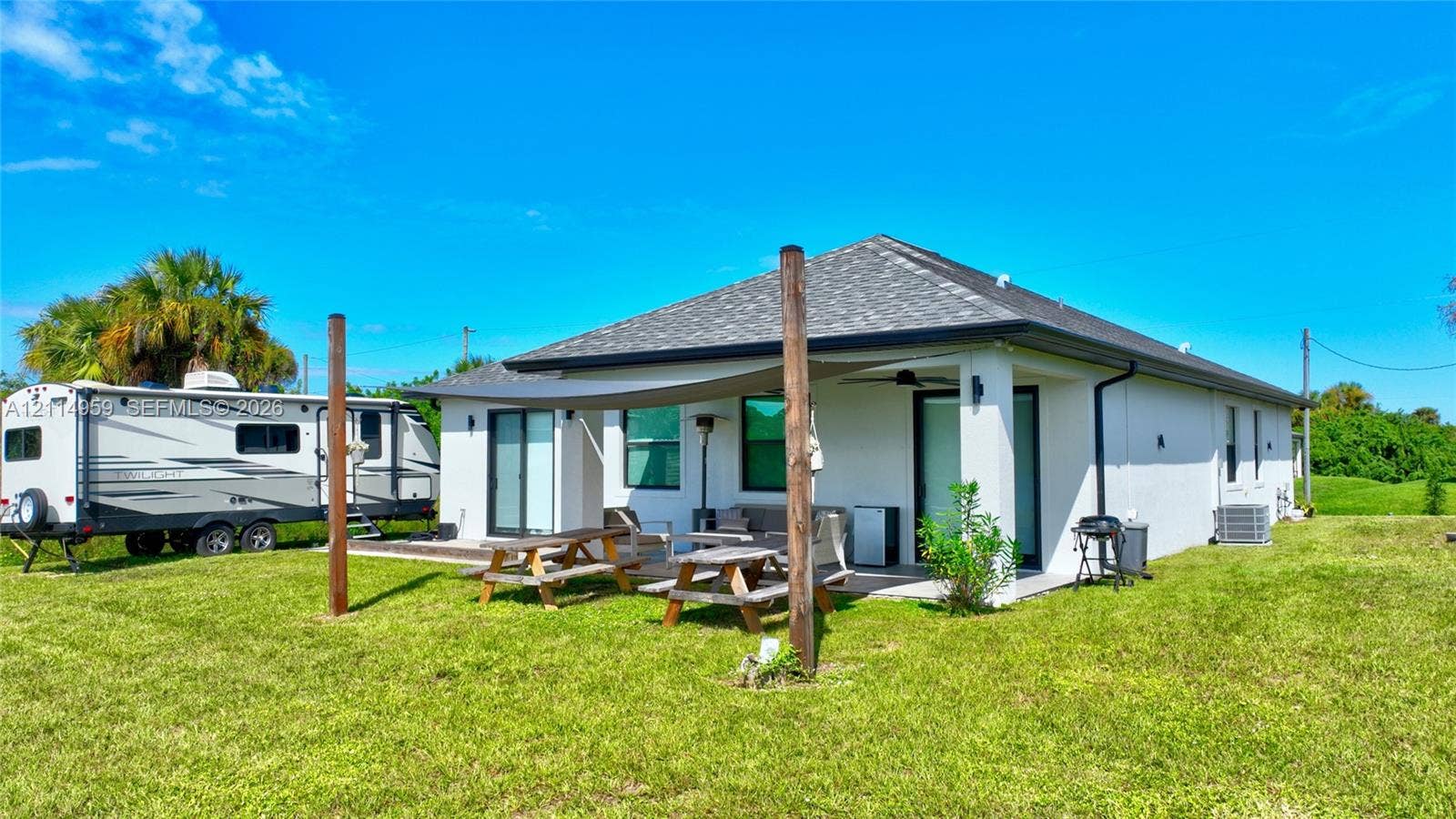 8017 Sunrise Circle, Labelle, FL 33935 Stucco exterior residence featuring a covered concrete patio, shaded outdoor area with fabric canopy, architectural shingle roof, and expansive lawn