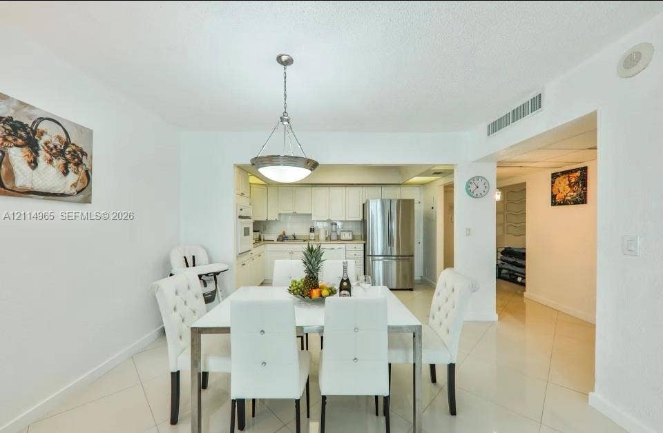 19370 Collins Avenue, #414, Sunny Isles Beach, FL 33160 Dining area featuring large-format ceramic tile flooring and a polished metal pendant light fixture, extending to a kitchen with white cabinetry and stainless steel refrigerator