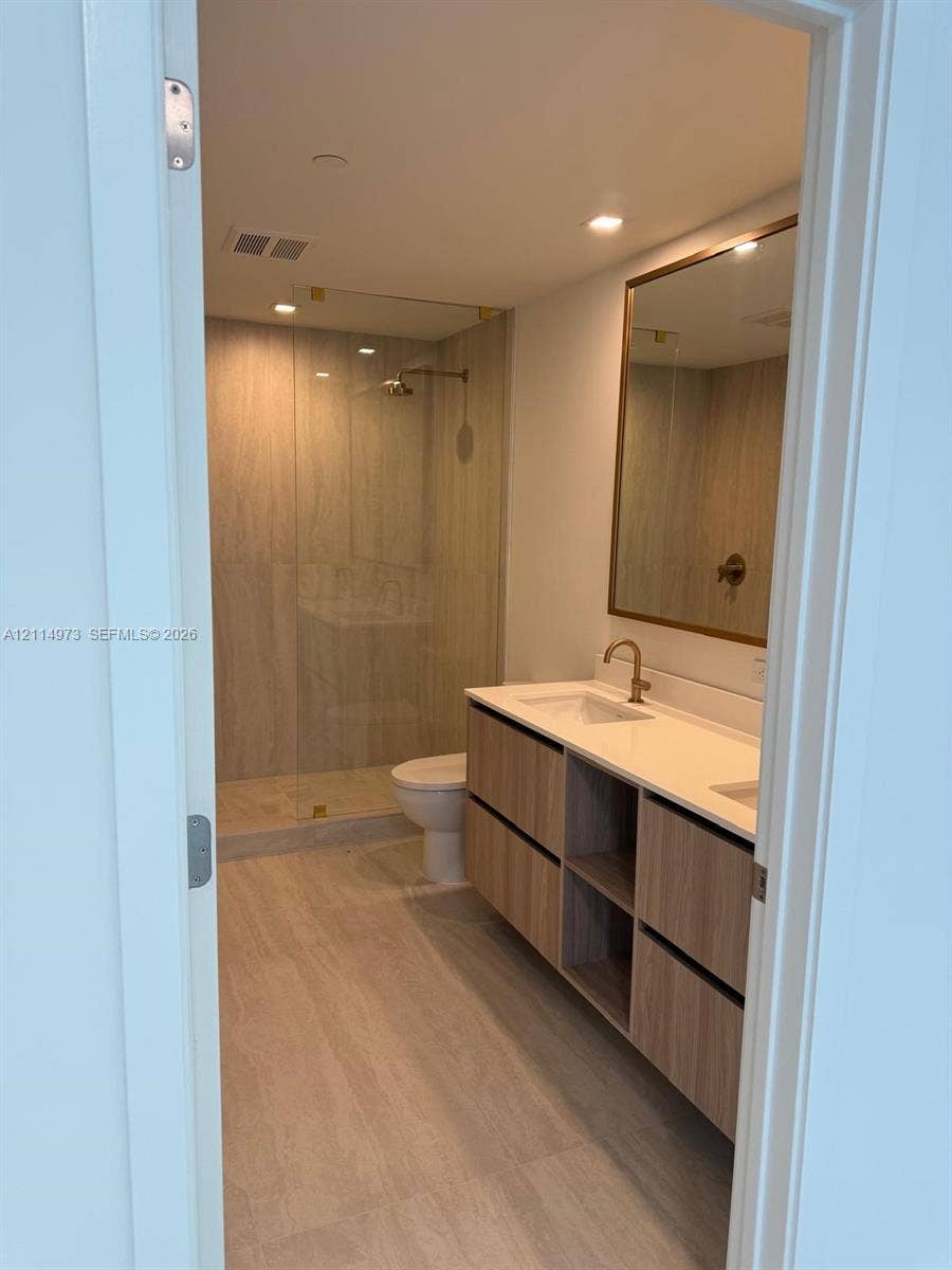 77 SE 5th Street, #2406, Miami, FL 33131 Bathroom featuring a floating vanity with wood-finish cabinetry, solid-surface countertop with dual under-mount sinks, gold-tone hardware, and a walk-in shower with floor-to-ceiling glass enclosure