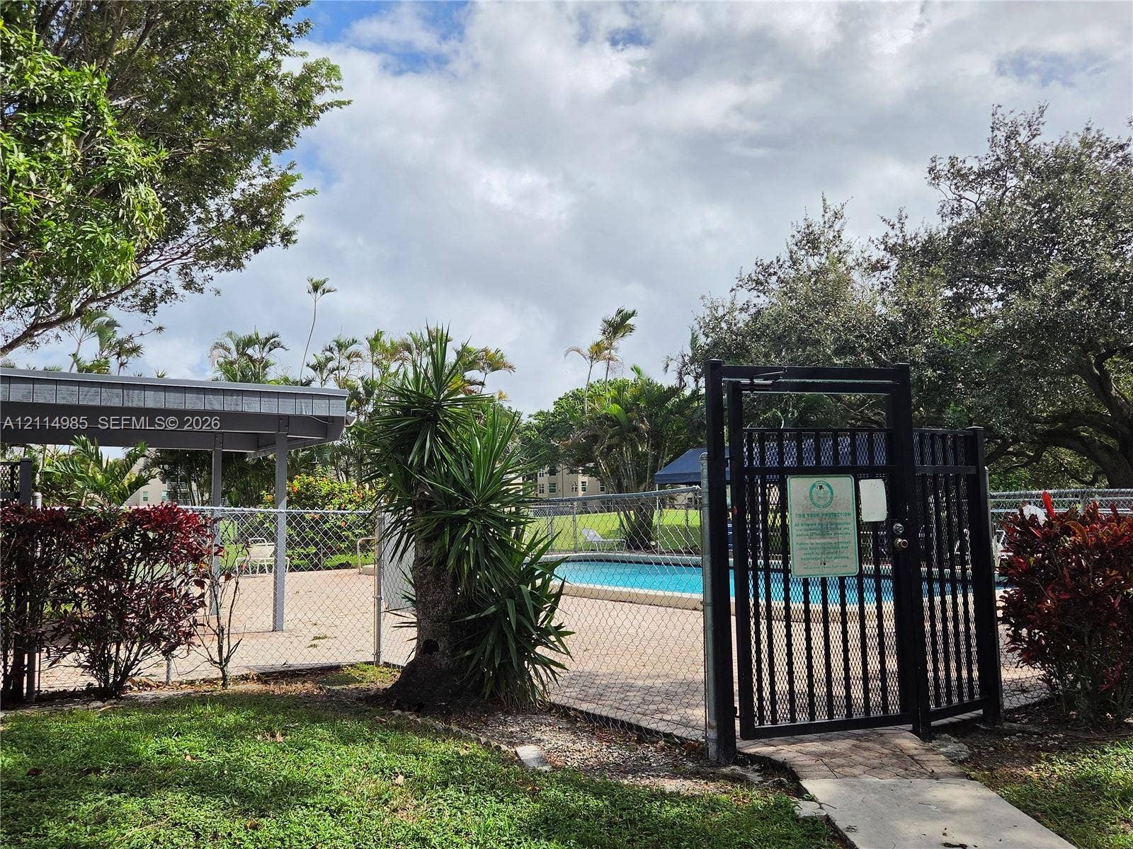 9441 Live Oak Place, #308, Davie, Davie, FL 33324 Outdoor swimming pool with paved decking, covered patio area, and metal security fencing surrounded by mature landscaping