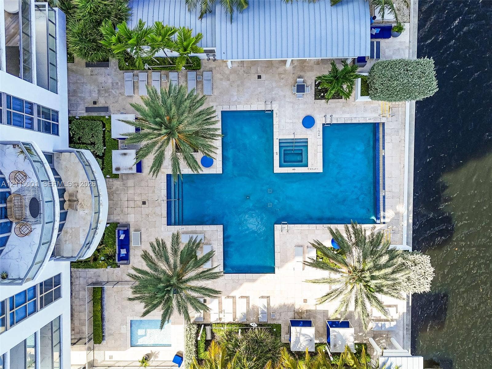 Waterfront swimming pool area featuring natural stone decking, integrated spa, mature palm landscaping, and metal-roofed covered terrace