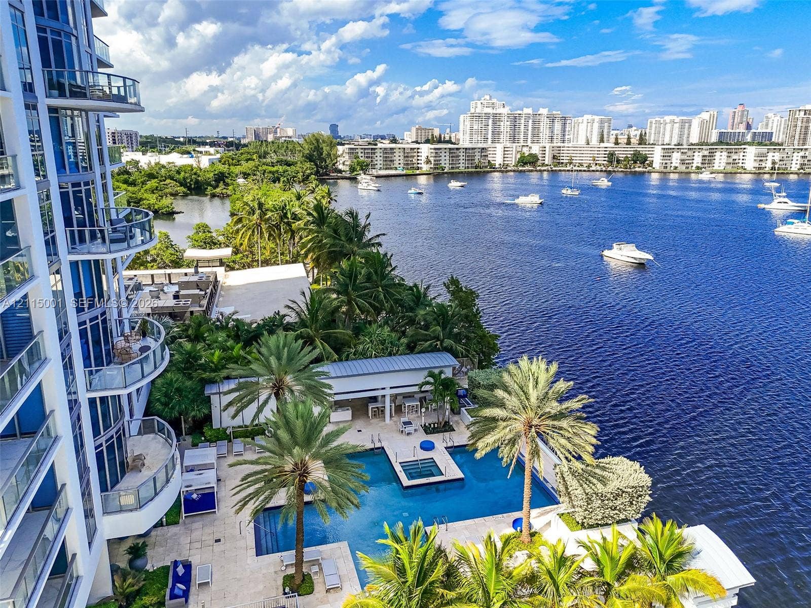 High-rise exterior featuring glass-railed balconies, waterfront swimming pool with integrated spa, stone-paved pool deck, and landscaped palm trees