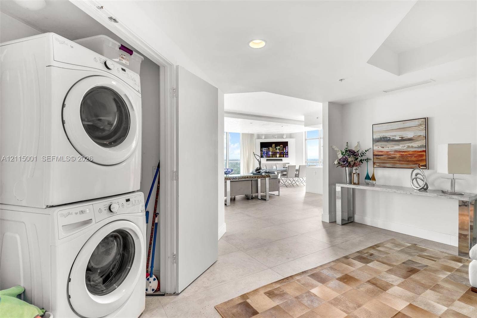 Stacked laundry unit featuring white front-load appliances, neutral large-format floor tile, and recessed ceiling lighting