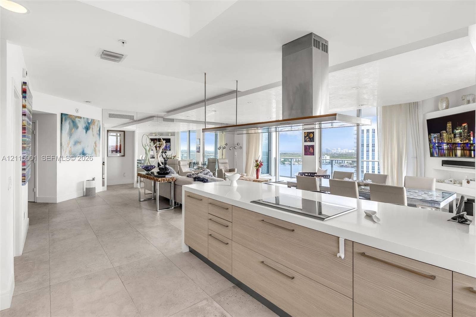 Open-concept kitchen featuring an expansive quartz-topped island, light-toned wood-finish cabinetry, a stainless steel chimney range hood, and tile flooring