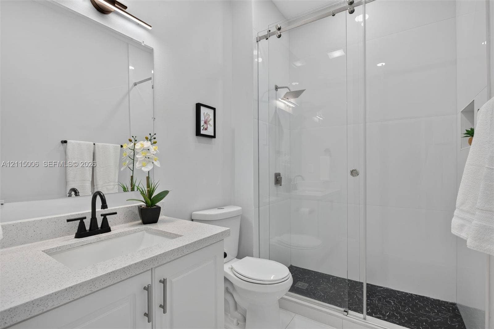 4812 NW 19th Street, Coconut Creek, FL 33063 White cabinetry vanity with solid-surface countertop, matte black faucet, glass-enclosed shower with contrasting dark stone-finish flooring, and wall-mounted light fixture