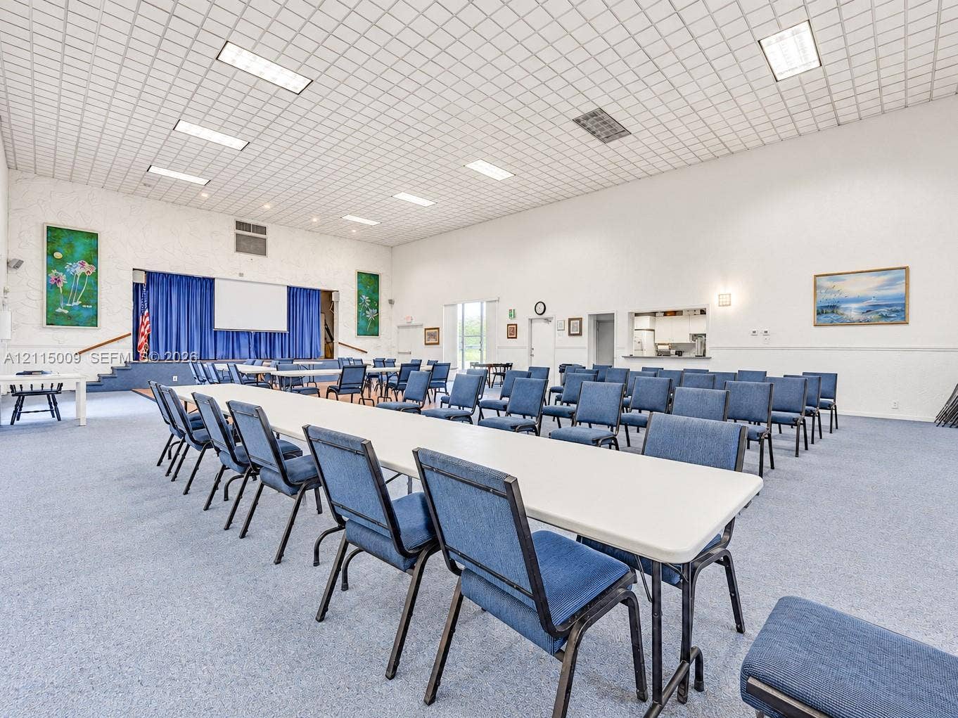 2300 Park Lane, #105, Hollywood, FL 33021 Expansive multi-purpose hall featuring a raised stage with curtain backdrop, recessed grid ceiling lighting, carpeted flooring, and a service pass-through window