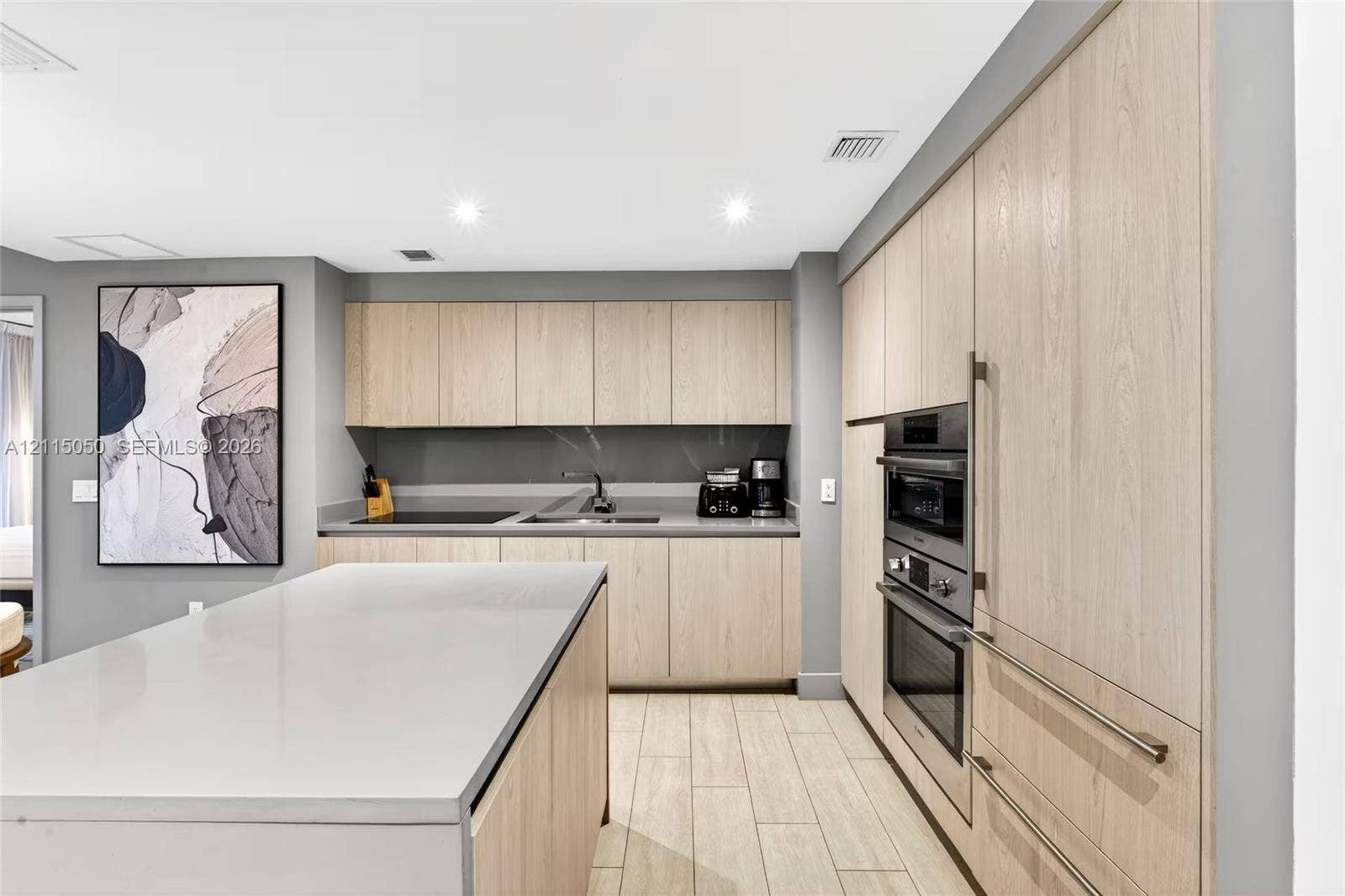 Kitchen featuring light-toned wood cabinetry, stone-surface island, built-in double wall oven, cooktop, and wood-finish flooring