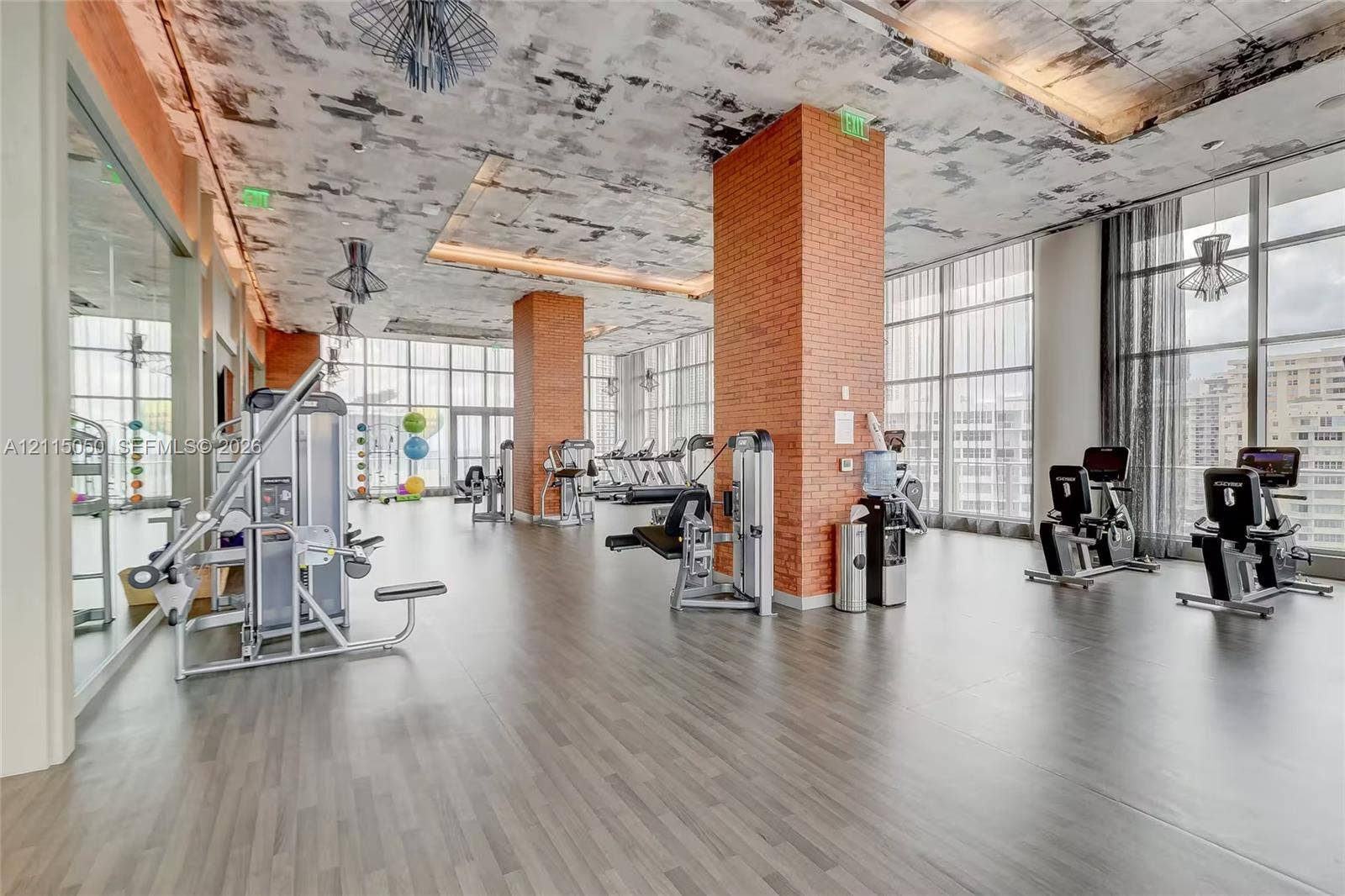 Fitness facility featuring floor-to-ceiling windows, exposed brick structural pillars, industrial-style concrete textured ceiling, and wood-finish flooring