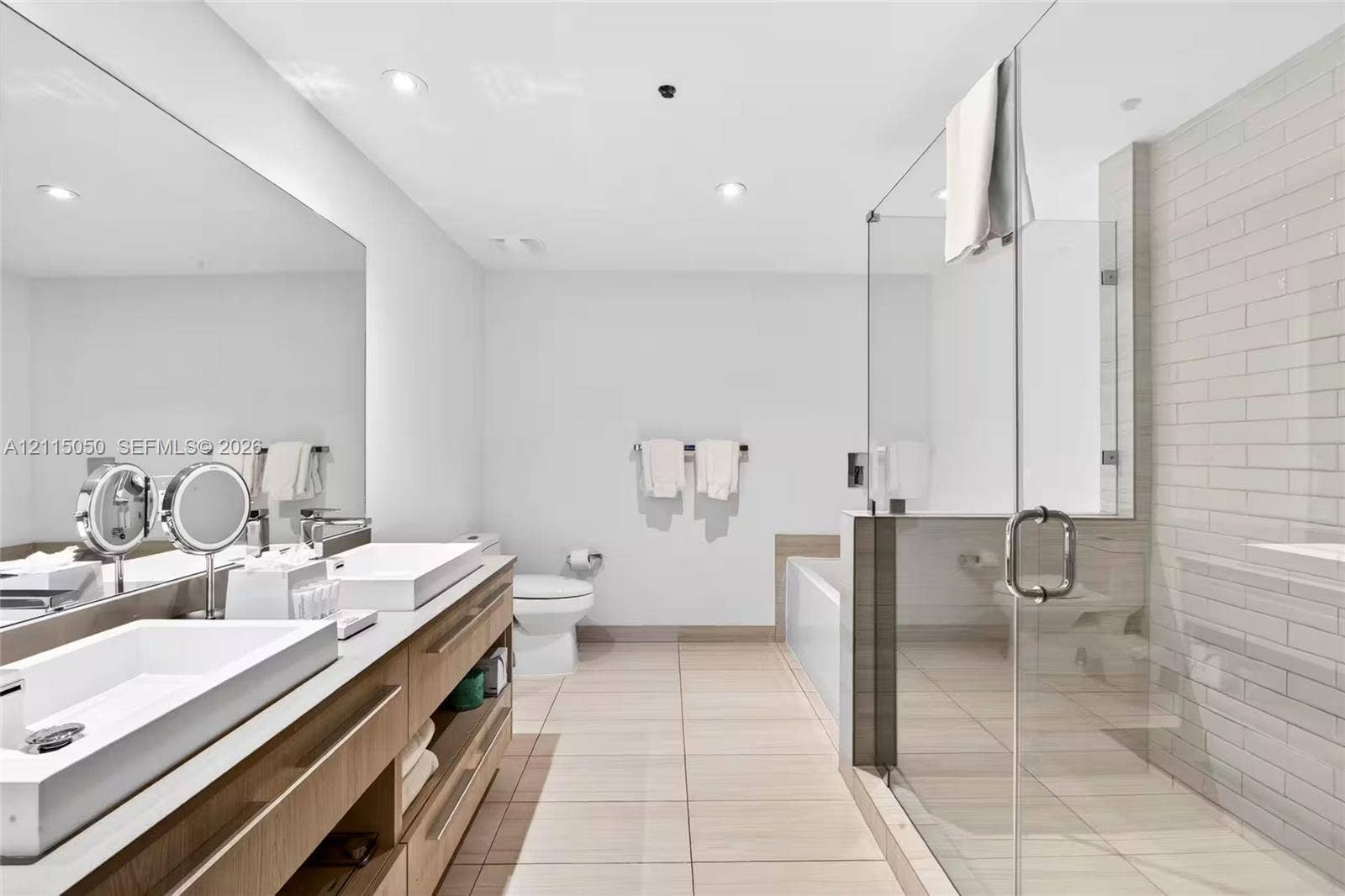 Dual-sink wood-finish vanity with rectangular vessel basins, frameless glass-enclosed shower, subway-style wall tile, and recessed lighting