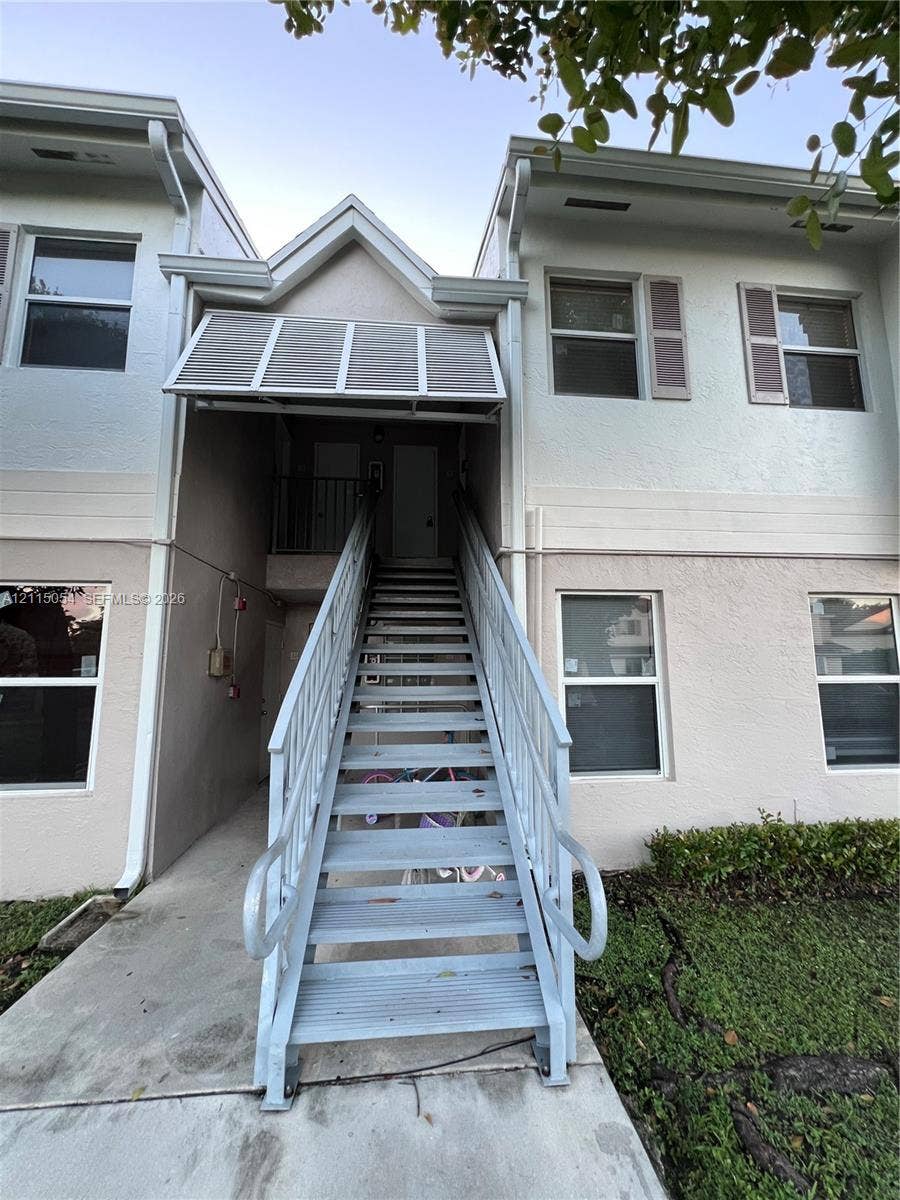 17500 NW 67th Court, #H-19, Hialeah, Hialeah, FL 33015 Two-story exterior featuring light-toned stucco siding, metal staircase with powder-coated railings, integrated window shutters, and a fixed metal awning over the entryway