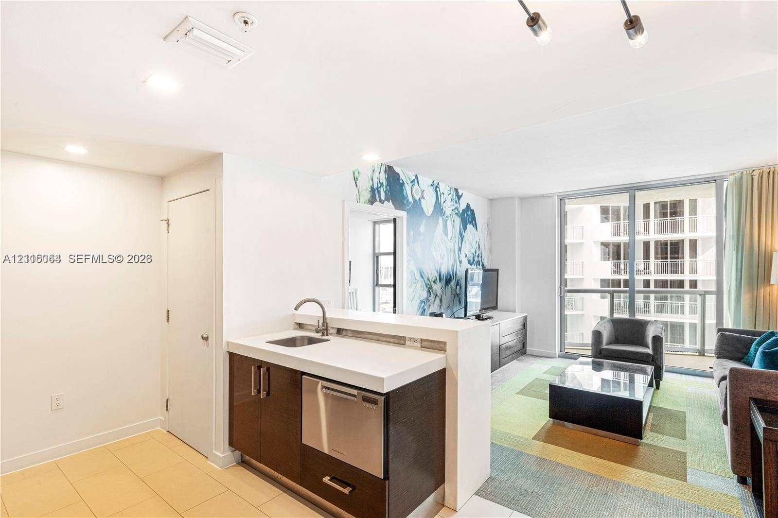 17315 Collins Avenue, #1808, Sunny Isles Beach, FL 33160 Kitchenette featuring dark cabinetry, solid-surface countertop, stainless steel undermount sink, and integrated stainless steel dishwasher, complemented by floor-to-ceiling glass panel windows