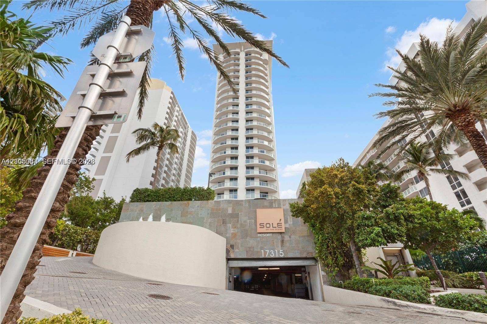 17315 Collins Avenue, #1808, Sunny Isles Beach, FL 33160 High-rise residential structure featuring a stone-clad facade, circular balcony design, and a paver-stone driveway leading to a covered parking garage entrance