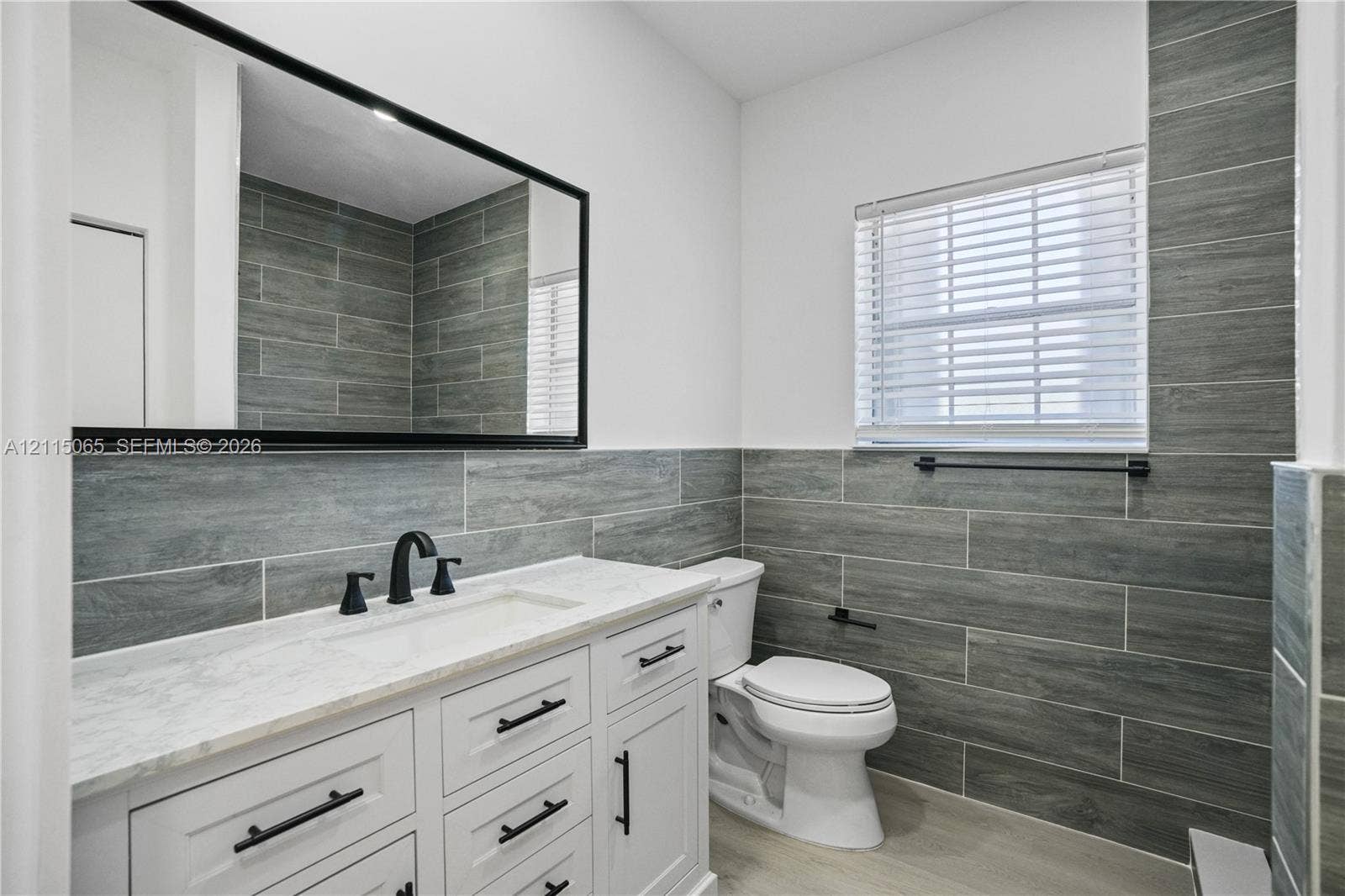 Bathroom vanity featuring white cabinetry, a stone countertop with an integrated rectangular sink, matte black hardware, and an expansive framed mirror, accented by horizontal grey stone-style wall tiling