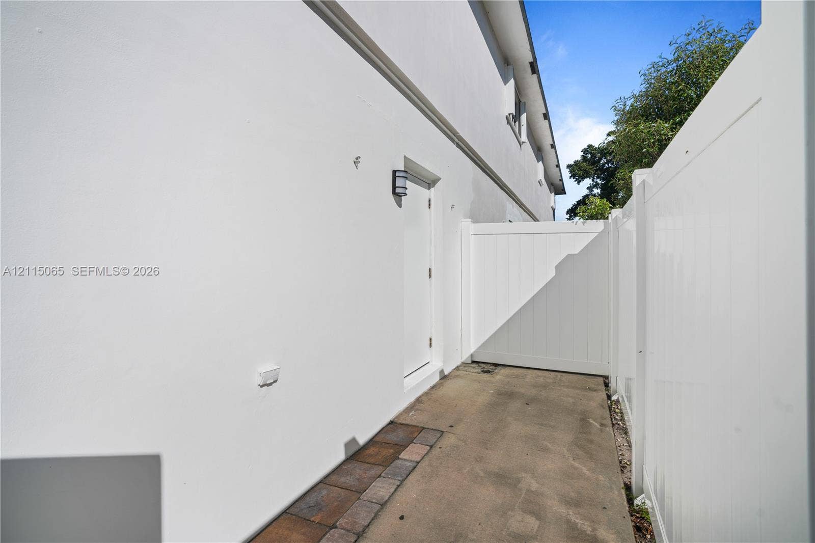 Exterior side entrance featuring a white solid-panel door, wall-mounted light fixture, concrete walkway with decorative stone paver border, and enclosed white vinyl fencing